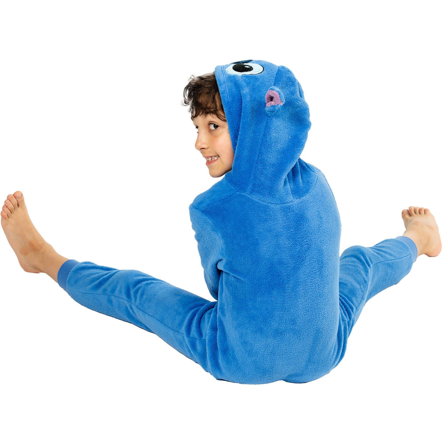 A child sitting playfully on the floor with legs spread apart, looking back over their shoulder while wearing a cozy blue Stitch onesie costume.