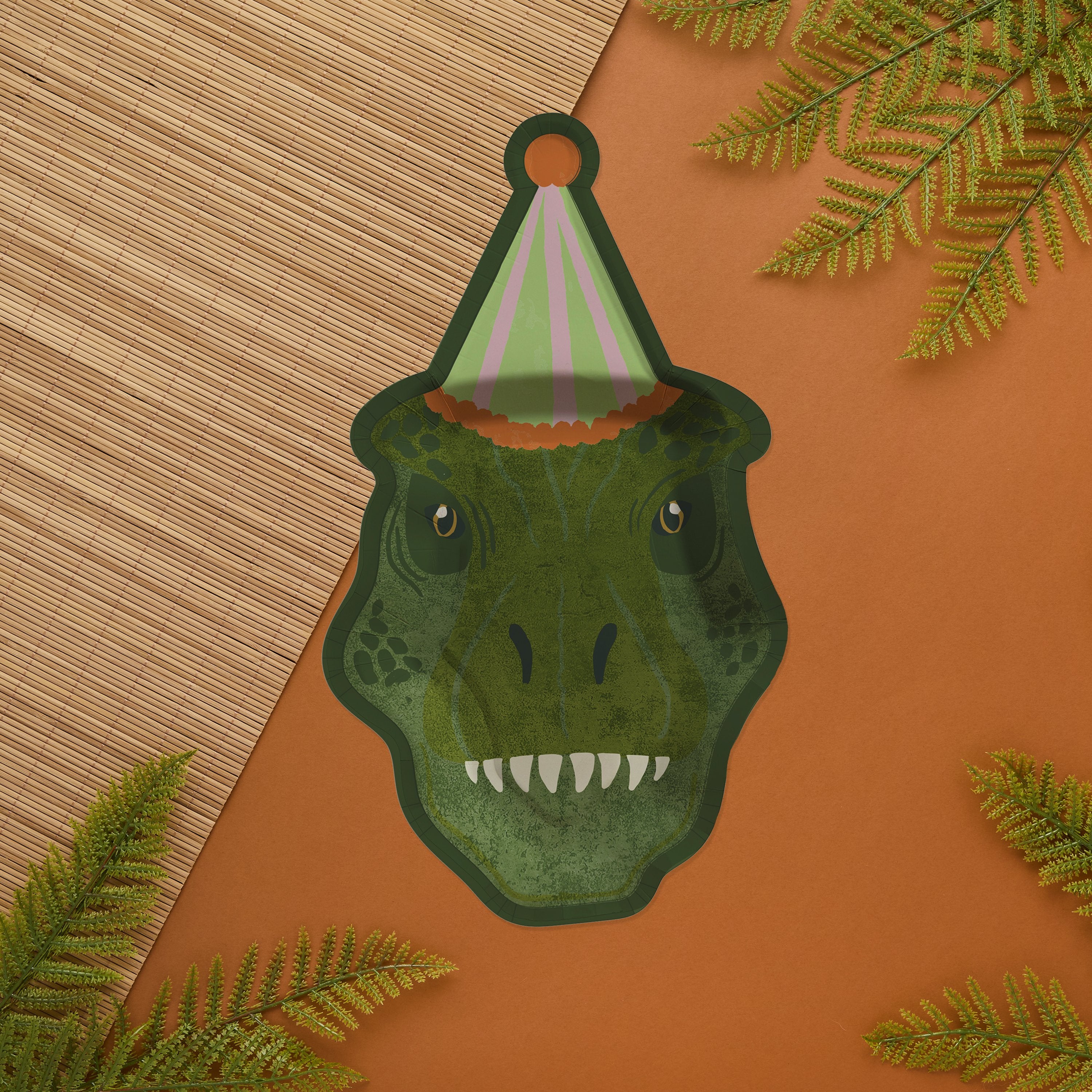 A T-Rex party plate on an orange background with bamboo matting and fern leaves, ideal for a themed birthday setup.