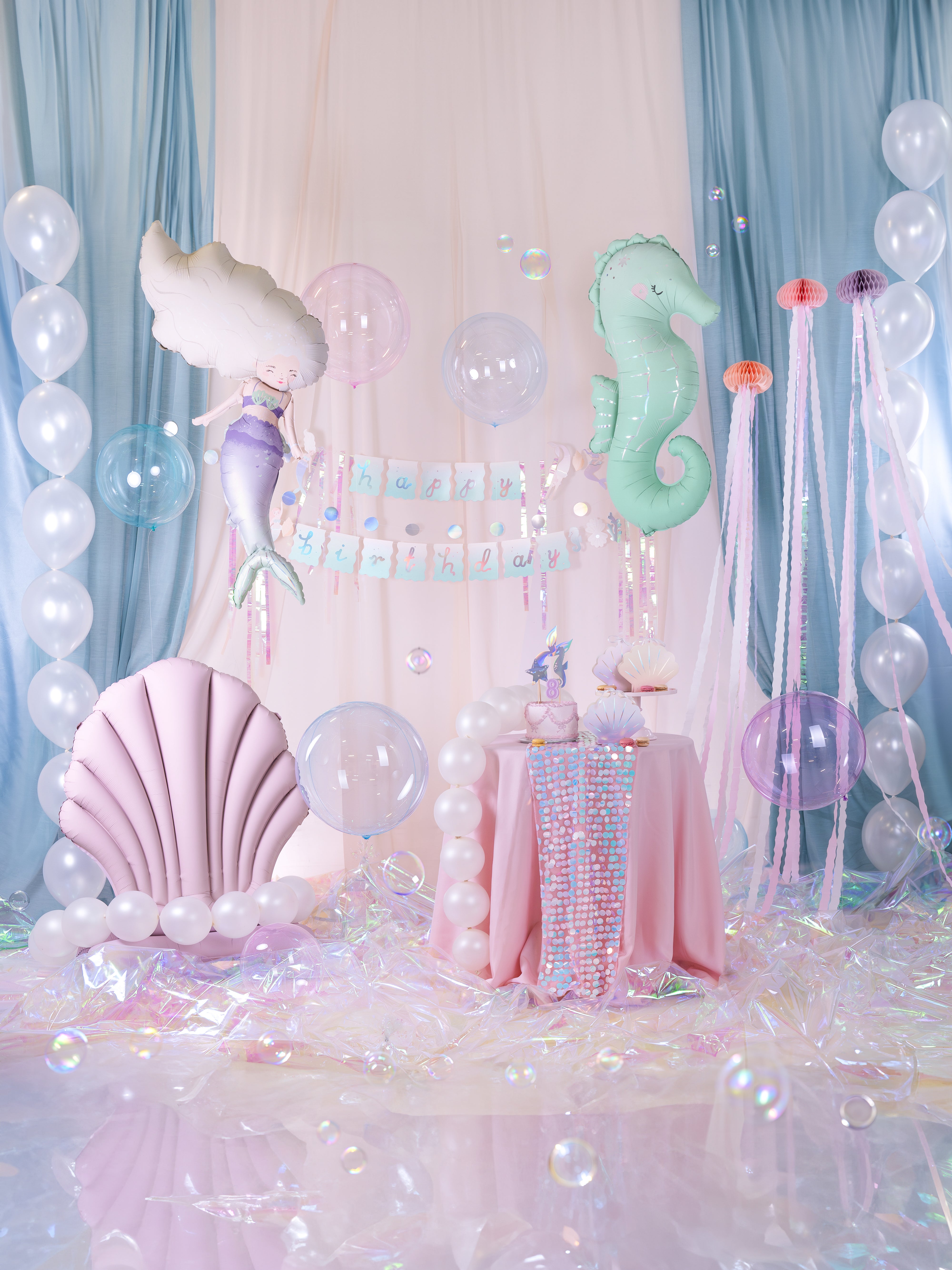 An elaborate mermaid-themed party setup with a draped backdrop of blue and white curtains, featuring a "Happy Birthday" banner, a large mermaid balloon, a large seahorse balloon, and columns of clear and white balloons. In the foreground, there's a pink seashell chair, a small round table with a pink tablecloth and iridescent runner holding a cake and other treats, an iridescent floor with bubbles, and several transparent pink sphere balloons floating among the decorations.