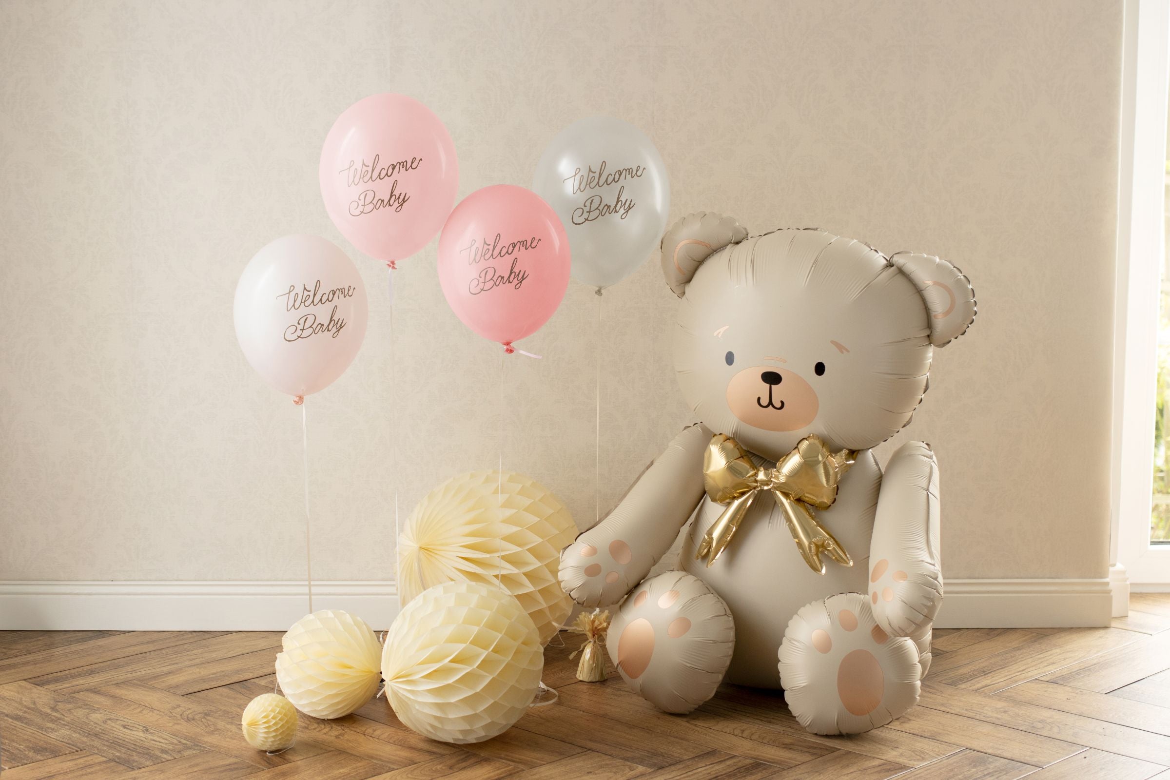 A cluster of four pink and white Welcome Baby balloons floating next to a large teddy bear balloon.