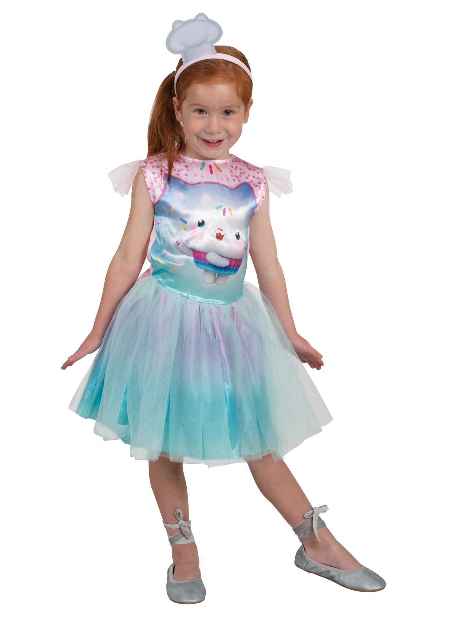 A red haired young girl modelling a pastel Gabby's Dollhouse Cakey Cat fancy dress costume, featuring a sprinkle-printed bodice of Cakey Cat with sheer flutter sleeves, an ombre purple and blue tulle tutu skirt, and a matching mini white chef hat headband.