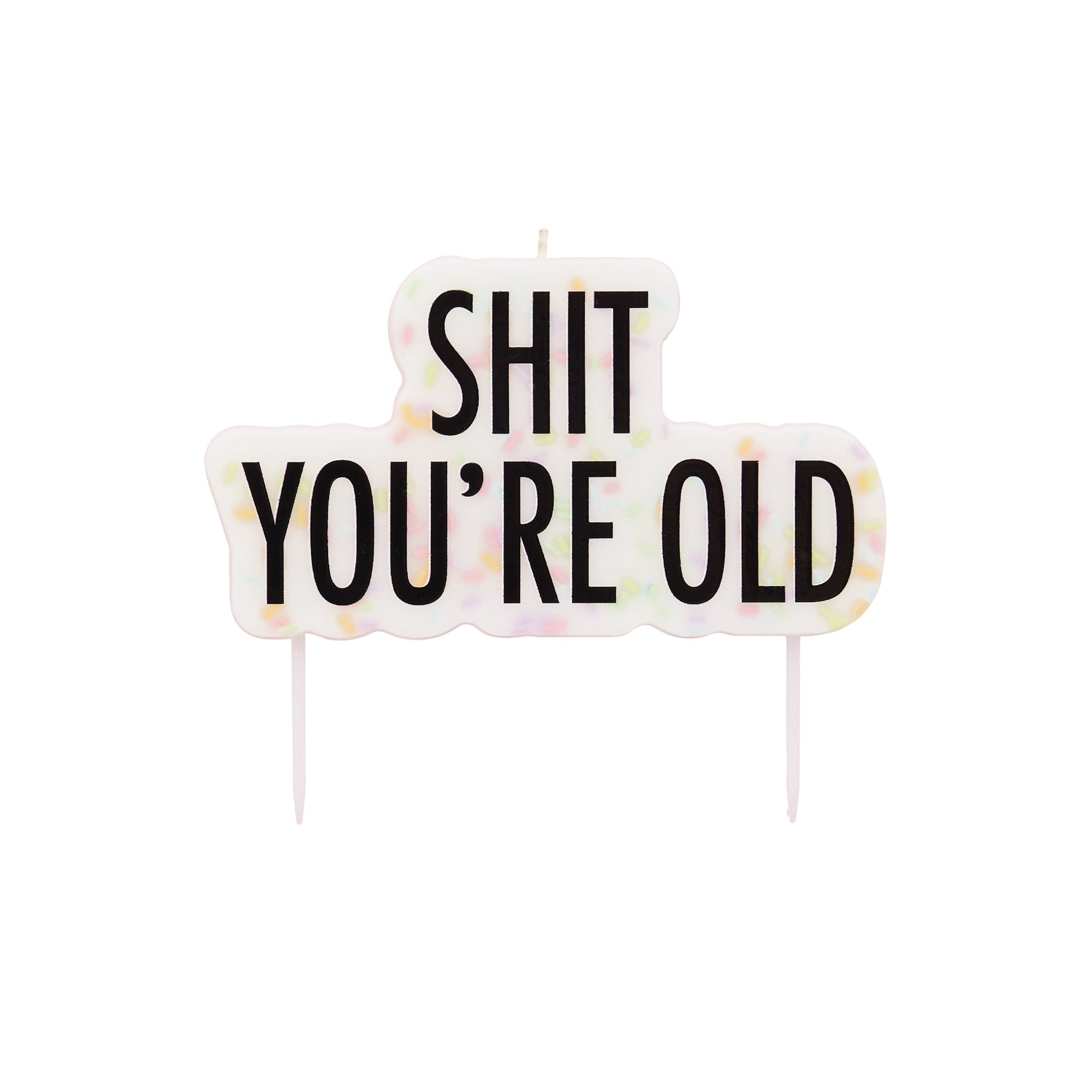 A pastel rainbow frosted birthday cake sitting on a white cake stand, topped with a lit novelty candle that reads "SHIT YOU'RE OLD" against a bright pink background.